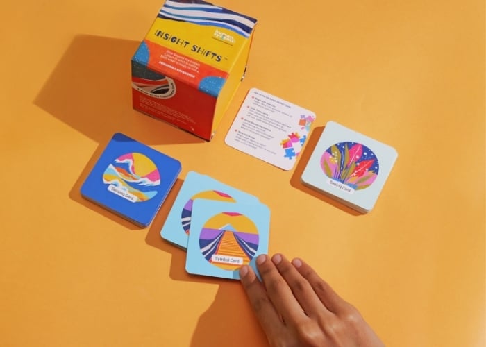 A hand selects a colorful card from a spread of illustrated cards labeled Insight Shifts. The cards and their box rest on an orange surface, alongside an instruction card.
