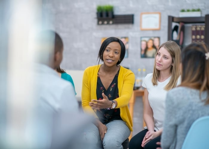 A woman in a yellow cardigan speaks with an open hand gesture while sitting in a group discussion with three other people in a cozy, informal setting.