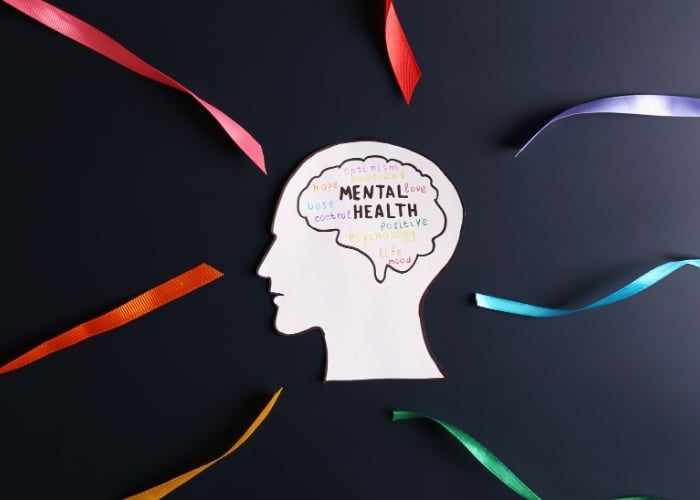 A paper cutout of a human head with words like mental health, hope, love, and optimism inside the brain. Colorful ribbons surround the head on a dark background.