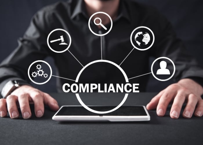 A person in a dark shirt sits at a table with a tablet, surrounded by compliance-related icons: a gavel, magnifying glass, globe, checklist, and user profile, linked to the word “COMPLIANCE.”.