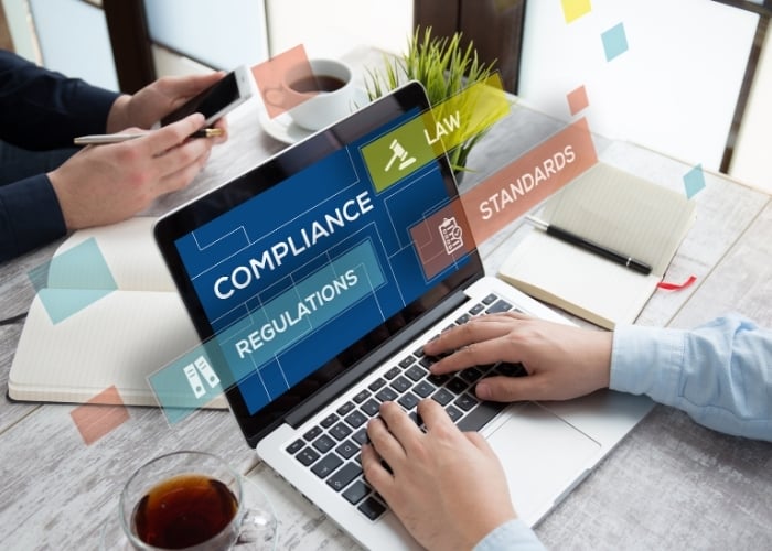 Two people work at a desk with a laptop displaying the words COMPLIANCE, REGULATIONS, LAW, STANDARDS. Papers, a notebook, a smartphone, and a cup of tea are also on the desk.