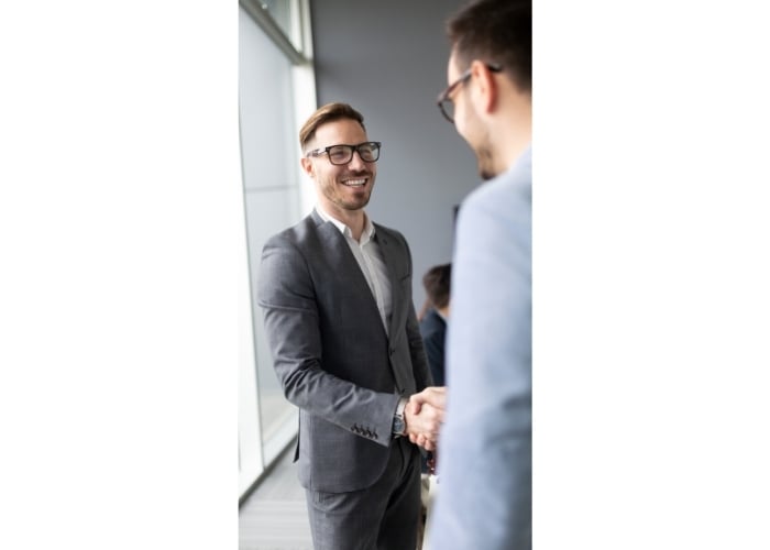 Two men in business suits shake hands in an office setting, smiling at each other. One man faces the camera, while the others back is turned. Natural light comes in from large windows.