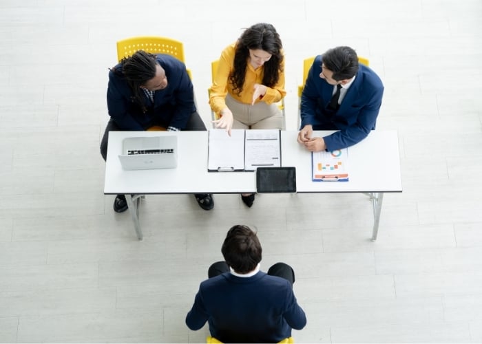 Four people sit around a white table in a meeting room. Three, dressed in business attire, face one person, suggesting a job interview or discussion. Papers, a laptop, and a tablet are on the table.