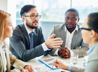 Four business professionals, two men and two women, sit at a table in a meeting room. One man is speaking while others listen attentively. Papers, charts, and a glass of water are on the table. Bright window light fills the room.