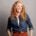 A smiling woman with long curly red hair wearing a denim shirt over a white top and rust-colored pants, standing against a plain gray background.