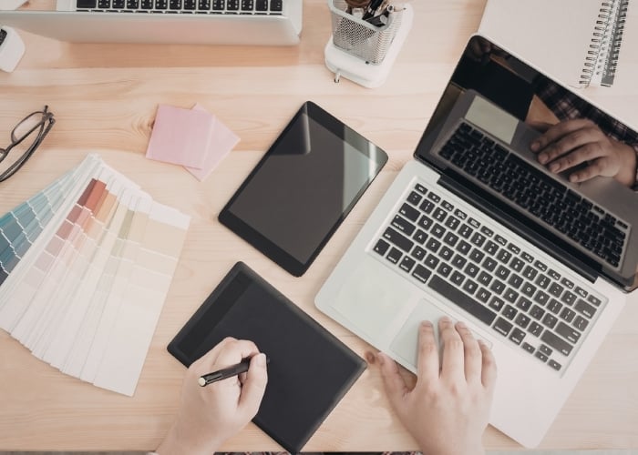 A person works at a desk with a laptop, tablet, and stylus, surrounded by color swatches, sticky notes, and office supplies, suggesting a digital design or creative workspace.