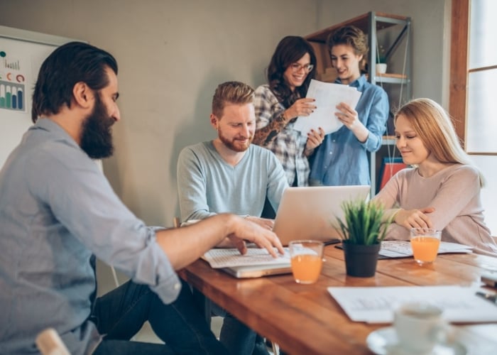 Four people sit around a table with a laptop, papers, and orange drinks, while two others stand behind them looking at documents, all appearing engaged in a collaborative work environment.