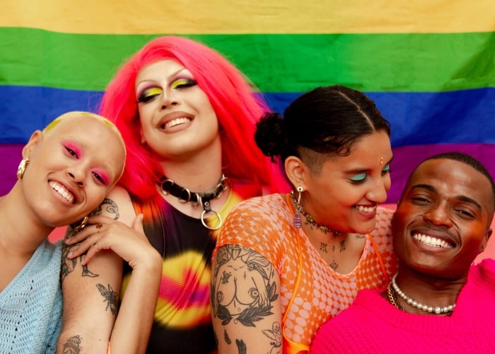 Four people with colorful makeup and hair smile and embrace in front of a vibrant rainbow pride flag, celebrating together and expressing joy and individuality.
