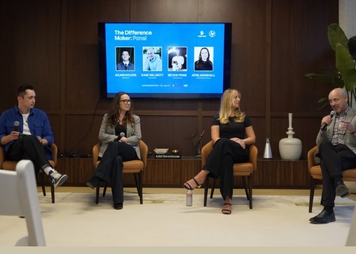 Four people sit on chairs in a panel discussion. A man on the right speaks into a microphone. Behind them is a screen displaying The Difference Maker: Panel with four headshots and names. The setting is modern and professional.