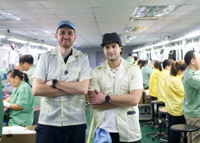 Two men in light uniforms and caps stand with arms crossed, smiling, in the foreground of a busy factory or assembly line where several workers in yellow uniforms are working at tables.