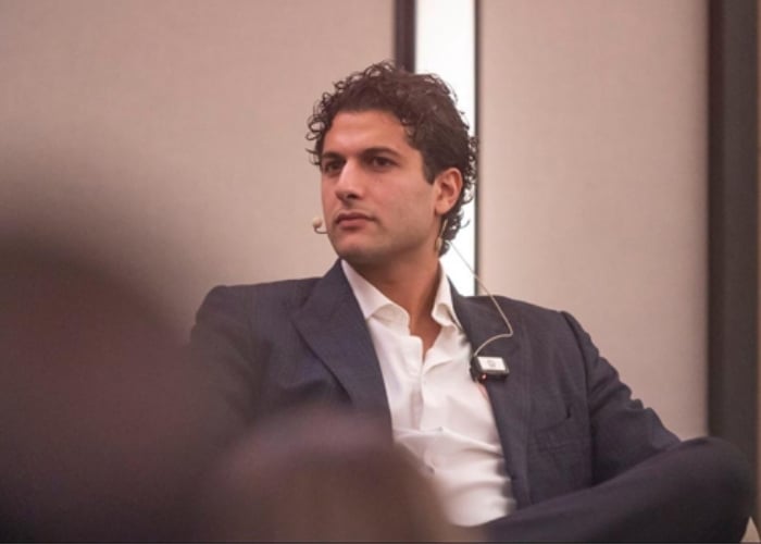 A man in a dark suit and white shirt sits, wearing a headset microphone, looking to the side in a conference setting with a neutral background.