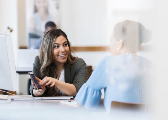 Two women sit at a table having a conversation. One woman, holding a smartphone and smiling, gestures toward the other. Papers and a laptop are on the table, suggesting a professional or business setting.