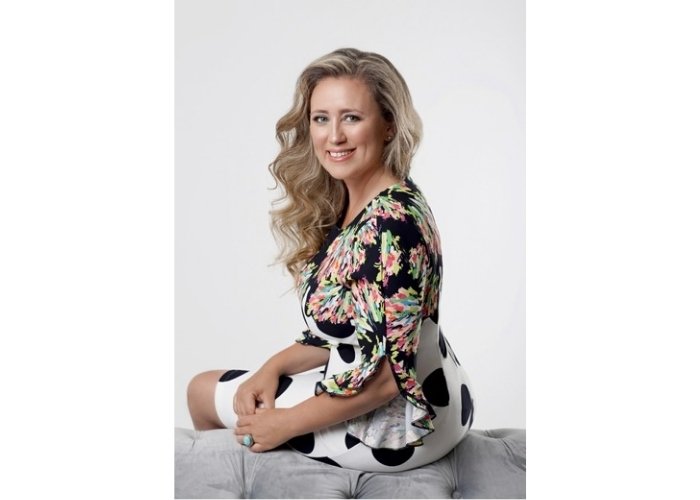 Smiling woman with long wavy blonde hair sits on a tufted gray ottoman wearing a floral top and white skirt with black polka dots.