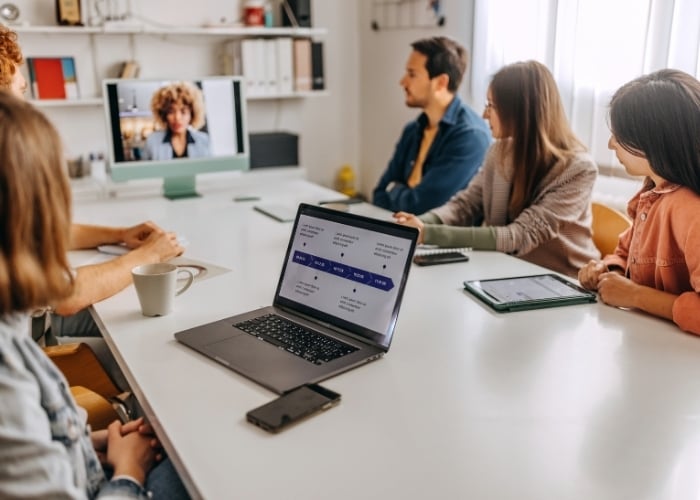 Five coworkers in a bright meeting room around a white table; a laptop shows slides and a remote participant appears on a screen.] ,