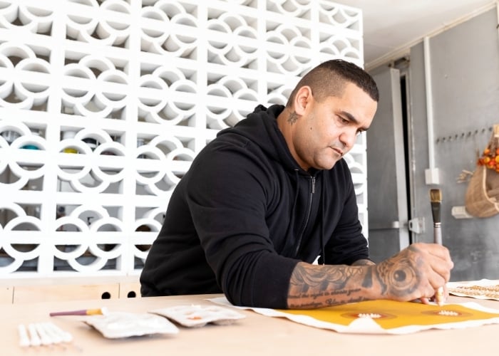 A man with tattoos, wearing a black hoodie, sits at a table painting on paper. Art supplies are scattered on the tabletop, and a white decorative wall is in the background.