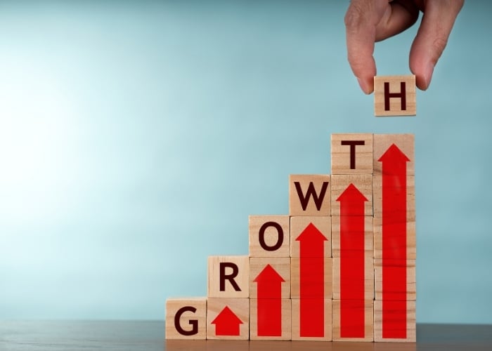 A hand places a wooden block with the letter H on top of a staircase of blocks spelling GROWTH, with red arrows pointing upward, symbolizing progress and development.