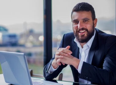 A man with a beard wearing a suit sits at a desk with a laptop, smiling and clasping his hands. A large window with a blurred outdoor view is in the background.