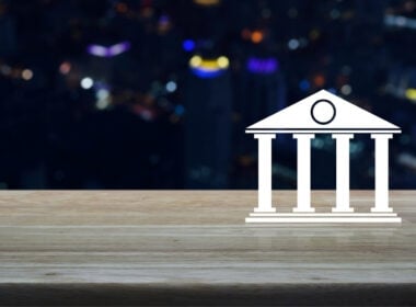 A white bank or courthouse icon is placed on a wooden surface, with a blurred cityscape featuring colorful lights in the background at night.