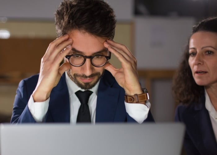 A man in a suit and glasses looks stressed, holding his head while staring at a laptop. A woman beside him appears concerned, watching him closely. Both are in an office setting.