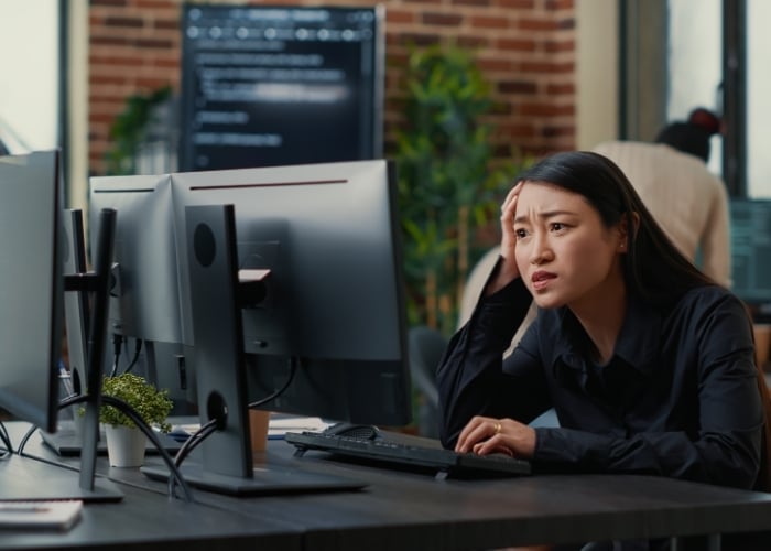 A woman sits at a desk with multiple computer monitors, looking frustrated and holding her head in one hand, in a modern office setting with brick walls and plants.