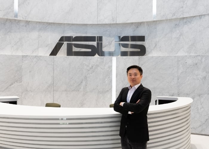 A man in a suit stands with arms crossed in front of a curved white reception desk with the ASUS logo on a marble wall behind him.
