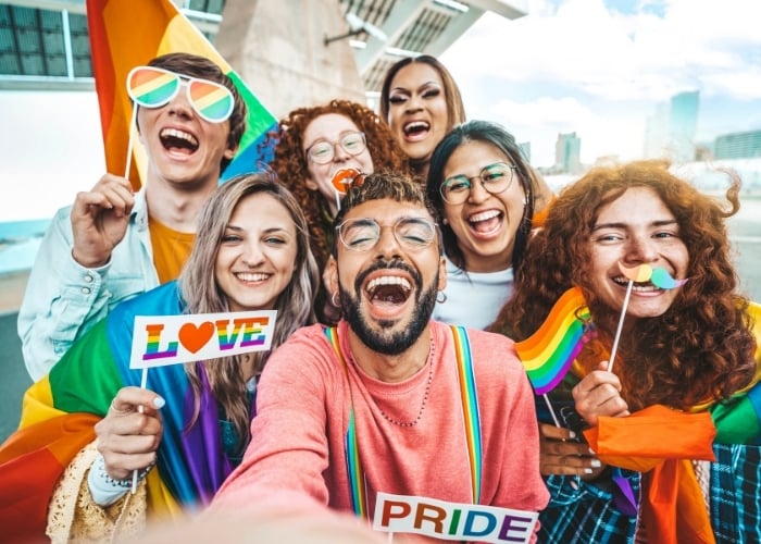 Diverse group posing for a Pride selfie, holding rainbow flags and signs saying LOVE and PRIDE.