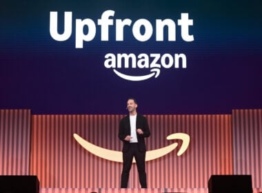 A person in business attire stands on stage in front of a large Amazon logo and the word Upfront displayed above. The background features vertical stripes in shades of orange and blue.
