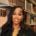 A young woman with long, straight black hair and a black jacket smiles while standing in a library, surrounded by shelves filled with books.