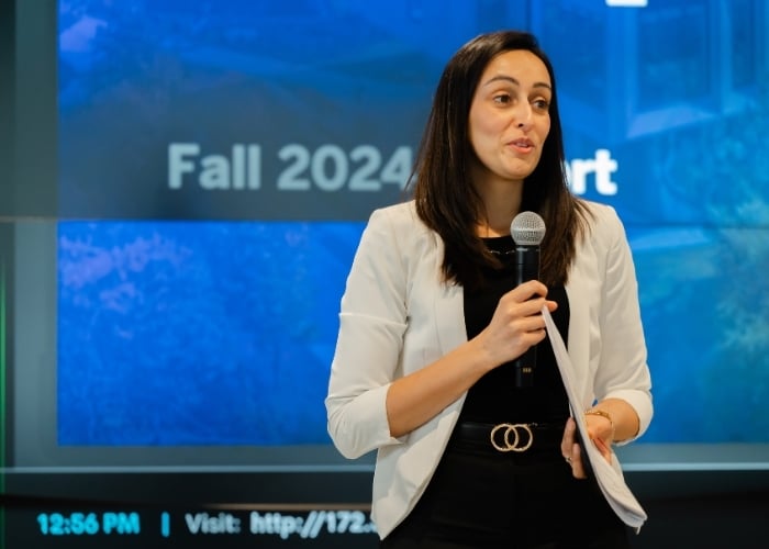 A woman in a white blazer holds a microphone and papers while speaking on stage. Behind her, a screen displays the words Fall 2024 and some blurred text and graphics.