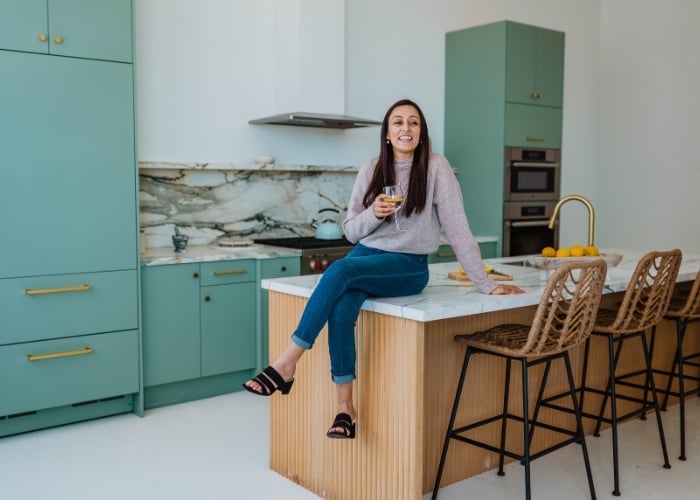 A woman sits on a kitchen island, smiling and holding a drink. The kitchen has teal cabinets, a marble backsplash, rattan bar stools, and a bowl of lemons on the counter.