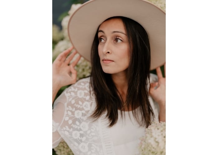 Woman with long brown hair wearing a wide-brimmed beige hat and a white lace top, looking to the side surrounded by light-colored flowers.