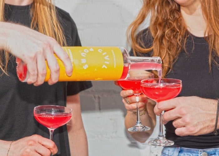 Two people hold coupe glasses with bright pink drinks as a third person pours more pink liquid from a yellow-labeled bottle. All are wearing black shirts and are standing against a light-colored background.