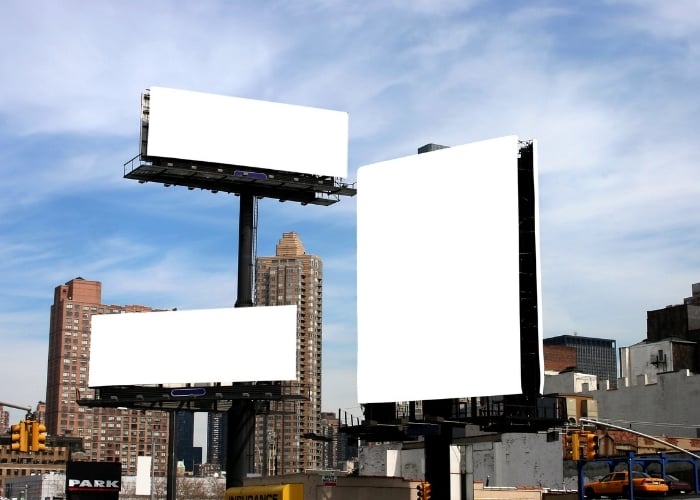 Three large blank billboards stand in an urban cityscape with tall buildings in the background and a blue sky with some clouds overhead.