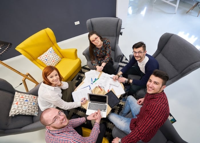 Five people sit in a circle of armchairs, smiling up at the camera, with papers, pens, and a laptop on a table in the center, suggesting a collaborative work or study session.