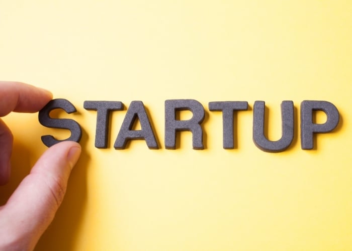 A hand arranges black letters spelling STARTUP on a bright yellow background.