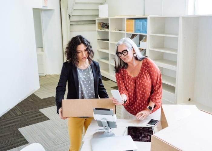 Two women standing at a counter in an office, one holding a cardboard box and the other scanning it with a smartphone; empty shelves and a staircase are visible in the background.