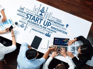 Four people sit around a table with laptops, tablets, and documents, discussing business. The table is covered with business-related words like START UP, innovation, strategy, and investment in various sizes.