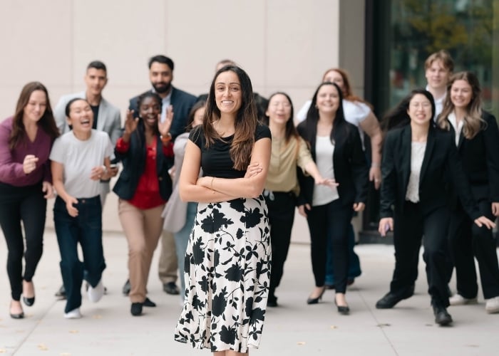 A woman stands confidently at the front with arms crossed, smiling, while a diverse group of people behind her walk forward enthusiastically, some also smiling and laughing, in an outdoor urban setting.