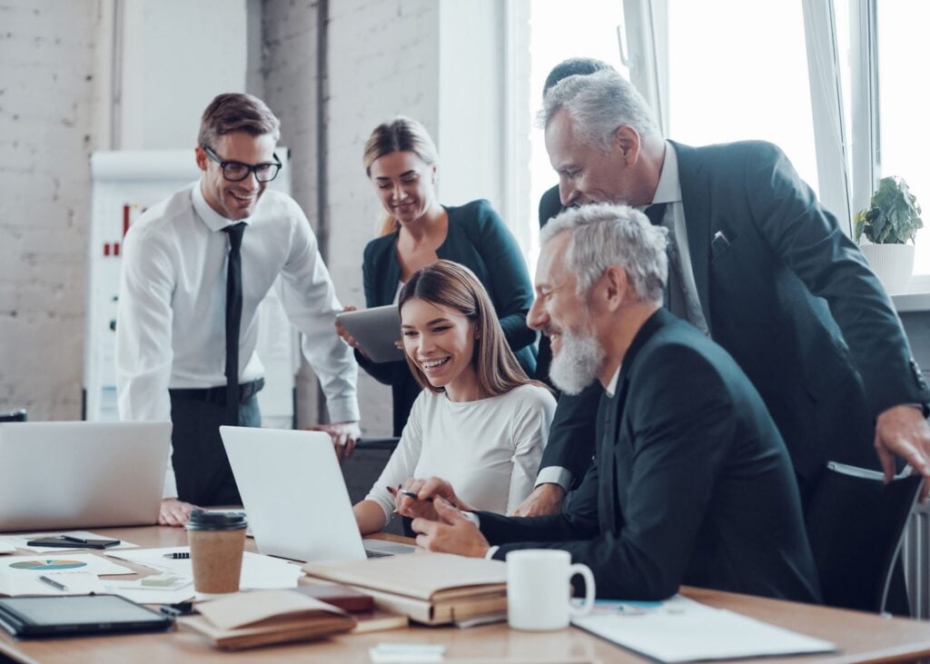 Five people in business attire gather around a desk, smiling and looking at a laptop screen, appearing engaged and collaborative in an office setting with papers and coffee cups on the table.