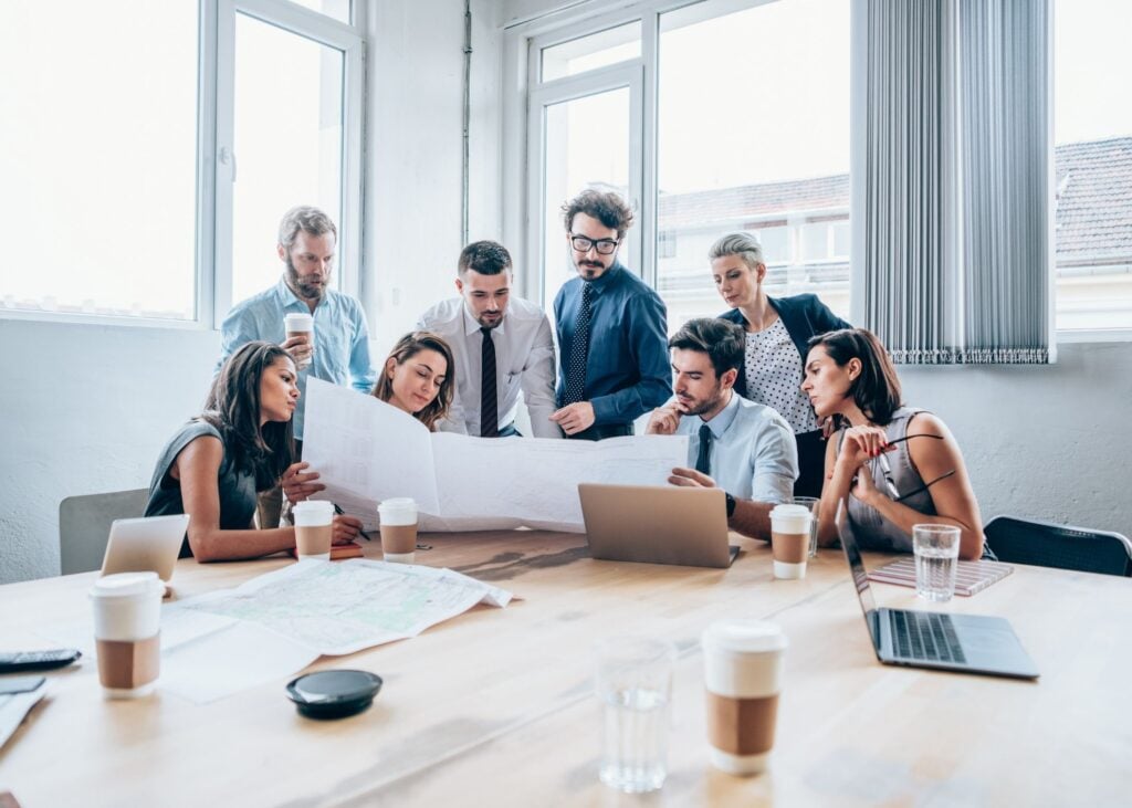Eight people in business attire gather around a table, examining large papers and working with laptops and coffee cups in a bright, modern office.
