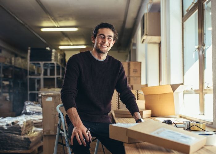 Smiling man sitting on a stool in a warehouse, surrounded by cardboard boxes, with sunlight coming through nearby windows.