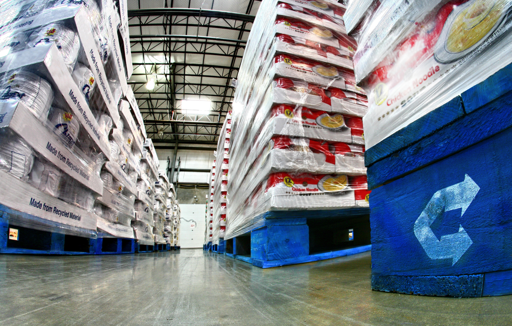 Stacks of shrink-wrapped goods on blue pallets are lined up in a warehouse aisle with a shiny concrete floor and high open ceiling, seen from a low angle.