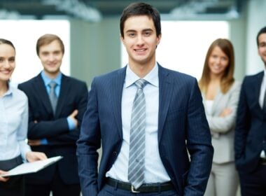 A young man in a suit stands confidently in front of four smiling colleagues dressed in business attire in an office setting.