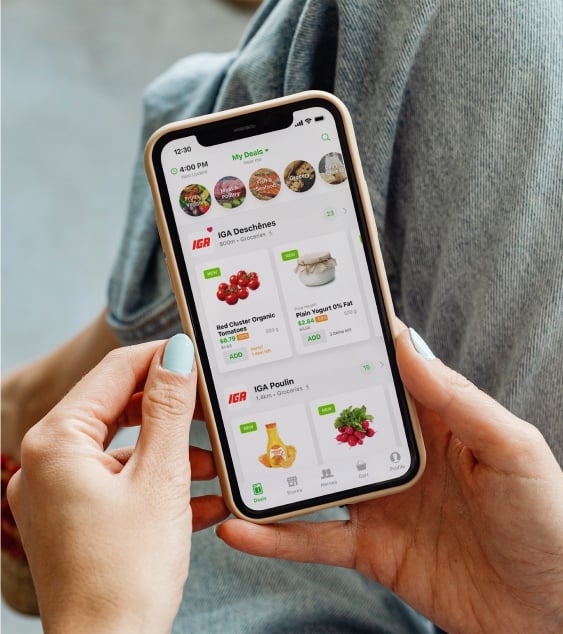 A person holding a smartphone with a grocery shopping app open, displaying deals on organic grapes, plain yogurt, juice, and beetroot. The user is seated, wearing jeans, with pale blue nail polish visible.