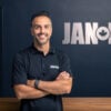 A man with dark hair and a beard, wearing a black JAN-PRO polo shirt, stands smiling with arms crossed in front of a wooden counter and JAN-PRO sign on a dark wall.